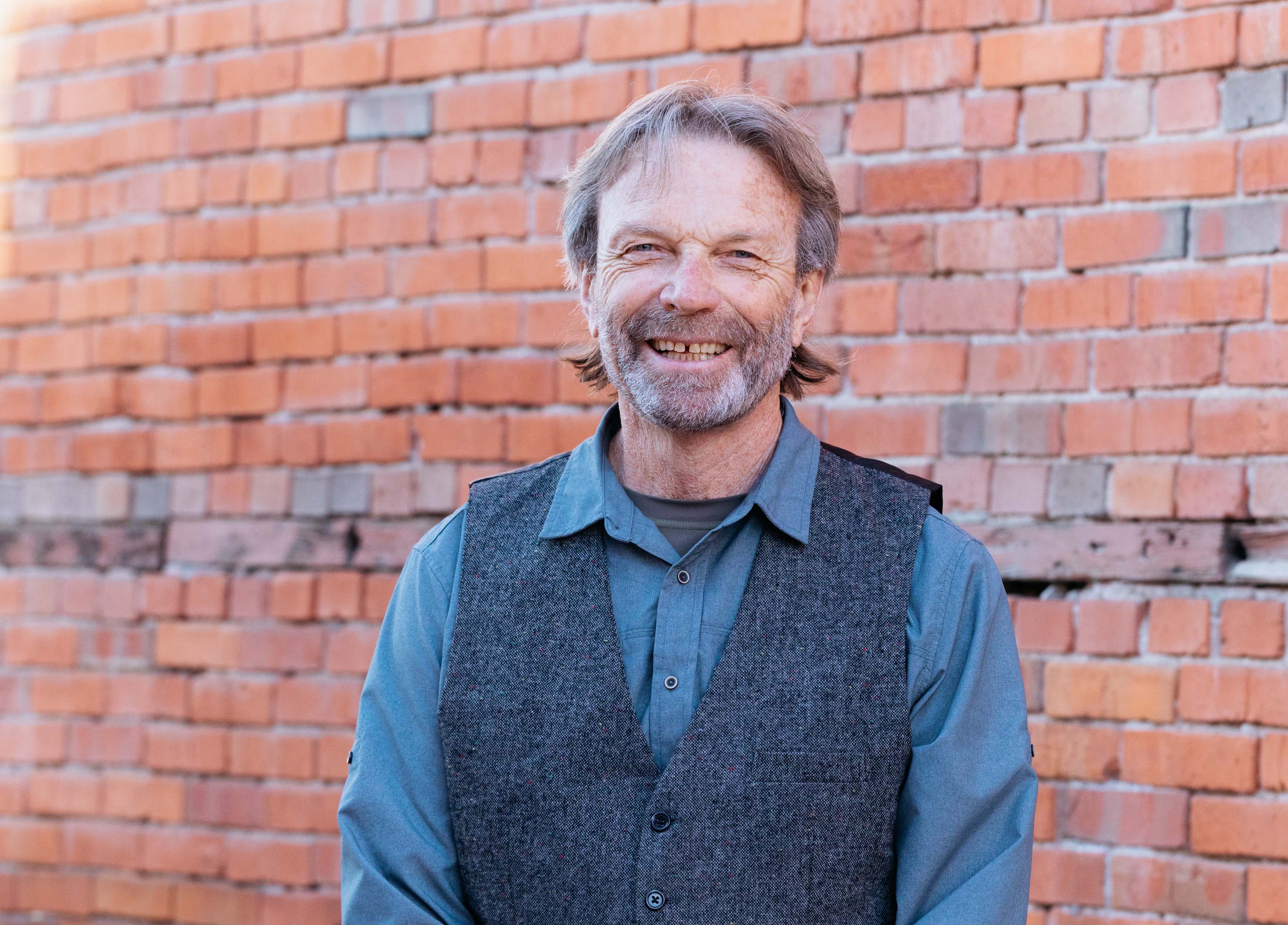 Glen Carmichael, middle aged man stands, smiling, in front of vintage brick wall in vest and blue collared shirt. 